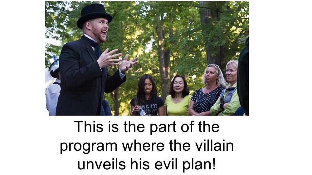 Image text: this is the part of the program where the villan unveils his evil plan! Image: interpreter wearing top hat and suit jacket speaking to visitors