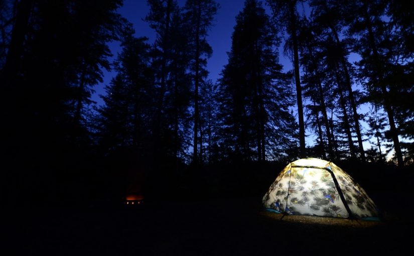 Emplacement de camping avec tente éclairée et petit feu qui brûle à la nuit tombée