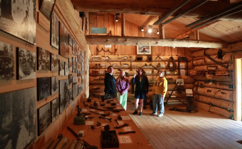 Un groupe de personnes visitant une exposition sur l'exploitation forestière à l'intérieur d'un bâtiment en rondins.