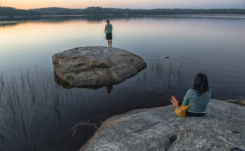 Une personne assise sur un rocher sur le rivage regardant une autre personne pêchant sur un rocher plus loin dans l'eau.