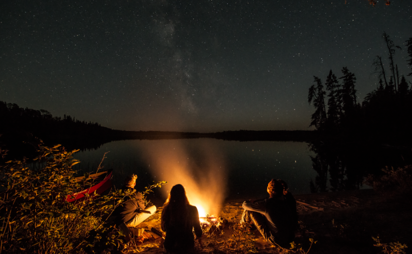 Group sits around campfire at night time by lake.