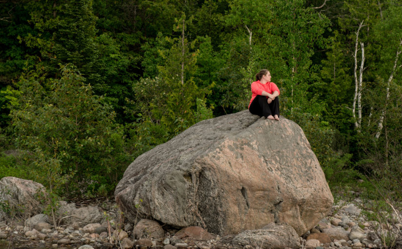 Woman sitting on a large boulder on the shore with a forest in the background