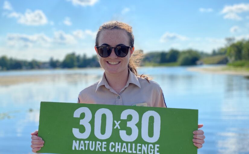 A person wearing sunglasses standing in front of a lake. They are holding a rectangular green sign that reads "30 x 30 Nature Challenge"